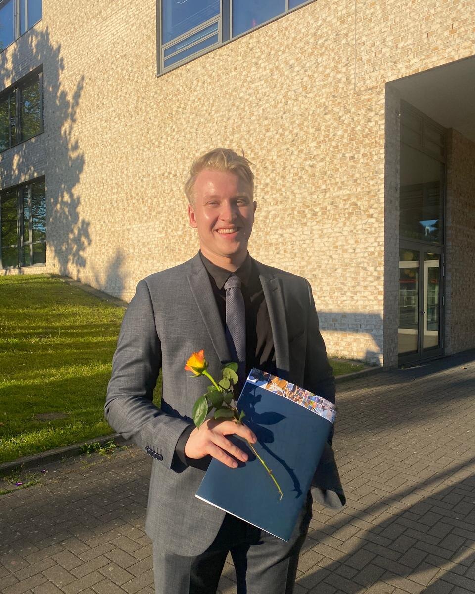 Finn Flebbe im Anzug mit Abiturzeugnis und Rose vor dem Schulgebäude